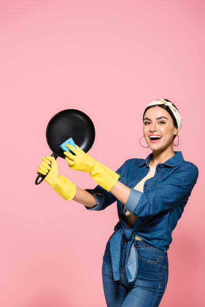 Gelukkig huisvrouw in rubber handschoenen houden koekenpan en spons geïsoleerd op roze  - Foto, afbeelding