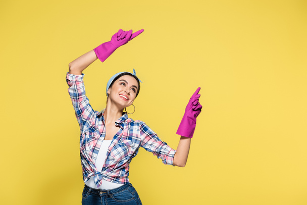Cheerful young housewife in rubber gloves pointing with fingers isolated on yellow  - Φωτογραφία, εικόνα