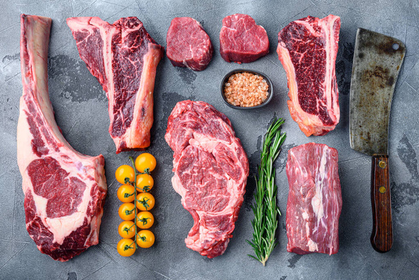 Különböző nyers marhahús steak grillezés fűszerezés és edények készlet, tomahawk, t csont, klub steak, borda szem és a szűzpecsenye darabok, szürke kő háttér, felülnézet lapos fektetés - Fotó, kép