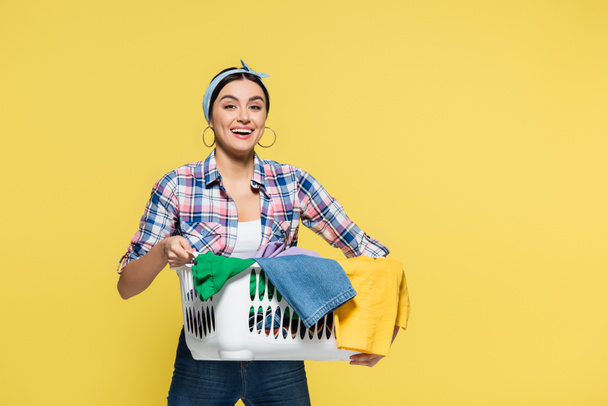 Young woman holding basket with clothes and smiling at camera isolated on yellow  - Foto, Bild