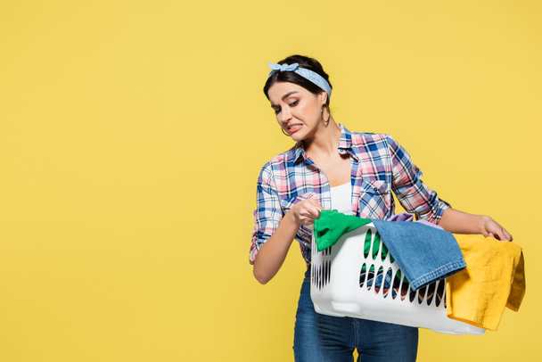 Confused housewife looking at clothes in basket isolated on yellow  - Photo, Image