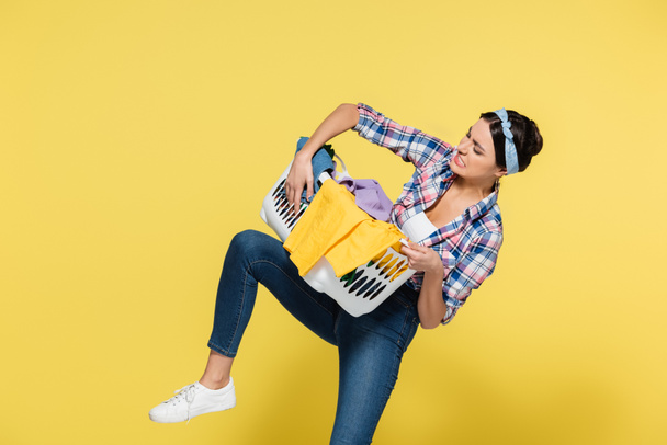 Angry woman holding basket with dirty clothes on yellow background  - Fotó, kép