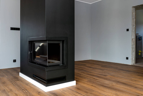 Ένα μοντέρνο τζάκι με κλειστό θάλαμο καύσης που στέκεται στο σαλόνι, βαμμένο μαύρο, με γωνιακό τζάμι καλυμμένο με αιθάλη. - Φωτογραφία, εικόνα