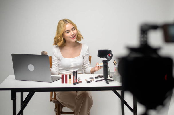 Behind the scenes young blonde woman entrepreneur working with laptop presents cosmetic products during online live stream over white background studio, selling online and beauty blogger concept - Photo, Image