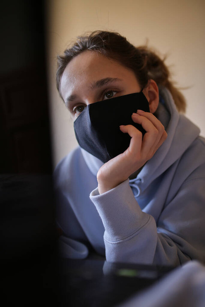 Ragazza caucasica con cappuccio blu e maschera nera funziona su laptop a casa isolato. Una ragazza con una maschera e una felpa blu lavora su un portatile a casa, seduta sul letto. Auto-isolamento. quarantena. COVID-19 - Foto, immagini