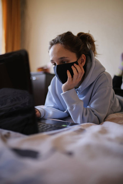 Biała dziewczyna w niebieskiej bluzie z kapturem i czarnej masce pracuje na laptopie w domu. Dziewczyna w masce i niebieska bluza pracują na laptopie w domu, siedząc na łóżku. Samoizolacja. kwarantanna. COVID-19 - Zdjęcie, obraz