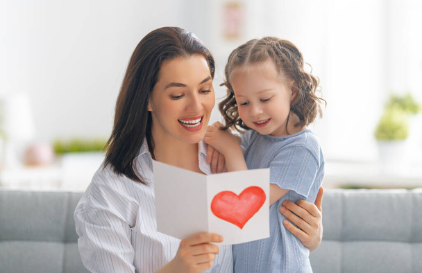 Happy mother's day! Child daughter is congratulating mom and giving her postcard. Mum and girl smiling and hugging. Family holiday and togetherness. - Фото, изображение