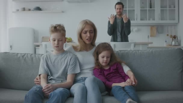 Δυστυχισμένη μητέρα να κάθεται στον καναπέ αγκαλιάζοντας θλιμμένα παιδιά και κλαίγοντας, θυμωμένος άντρας να φωνάζει και να μαλώνει στο παρασκήνιο - Πλάνα, βίντεο
