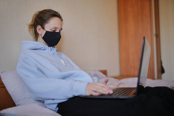 Biała dziewczyna w niebieskiej bluzie z kapturem i czarnej masce pracuje na laptopie w domu. Dziewczyna w masce i niebieska bluza pracują na laptopie w domu, siedząc na łóżku. Samoizolacja. kwarantanna. COVID-19 - Zdjęcie, obraz