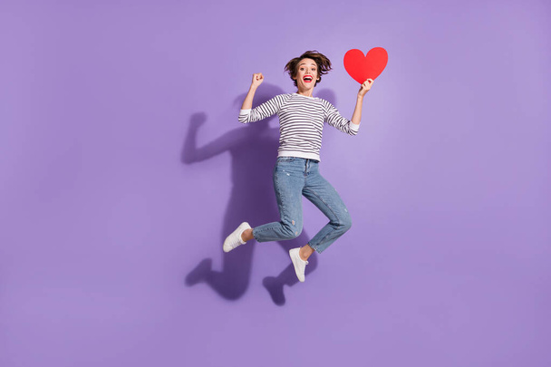 Full length photo of lovely young woman wear casual outfit jumping holding looking red paper heart isolated violet color background - Fotó, kép