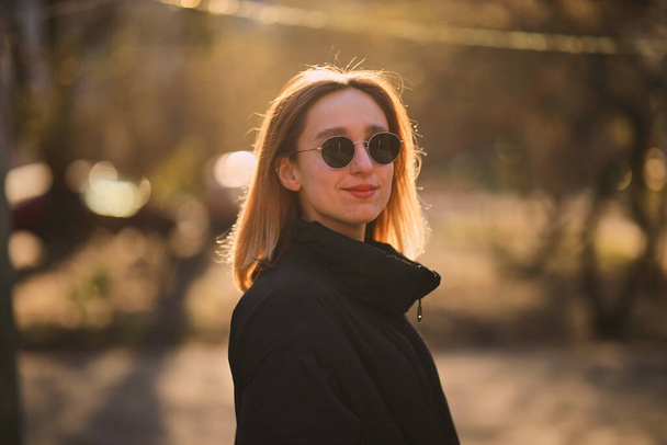 A blonde girl in a black jacket and black sunglasses walks on the street. beautiful girl in a black jacket and black glasses on the street. soft focus. - Foto, Bild