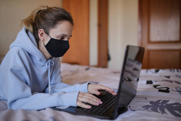 Caucasian girl in blue hoodie and black mask works on laptop at home isolated. A girl in a mask and a blue hoodie works on a laptop at home, sitting on the bed. Self-isolation. quarantine. COVID-19 - Foto, imagen