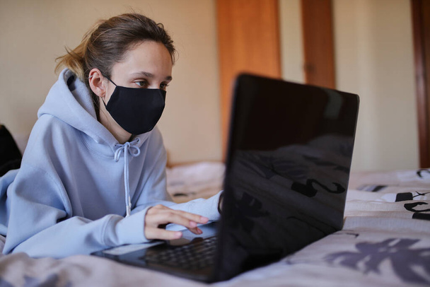 Caucasian girl in blue hoodie and black mask works on laptop at home isolated. A girl in a mask and a blue hoodie works on a laptop at home, sitting on the bed. Self-isolation. quarantine. COVID-19 - Fotó, kép