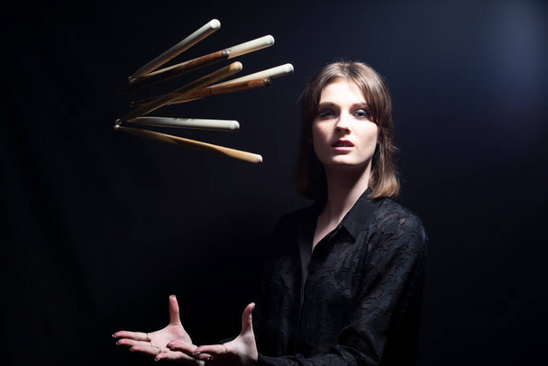 smiling drummer woman with drum sticks young beautiful musician girl tossing up effectively drum sticks. Dramatic portrait on black background - 写真・画像