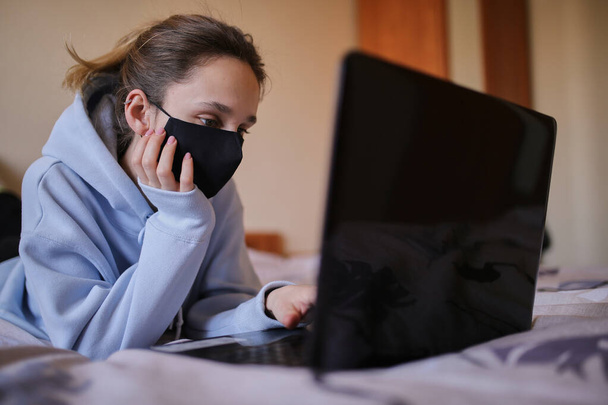 Menina caucasiana com capuz azul e máscara preta funciona no laptop em casa isolado. Uma menina com uma máscara e um capuz azul trabalha em um laptop em casa, sentado na cama. Auto-isolamento. quarentena. COVID-19 - Foto, Imagem