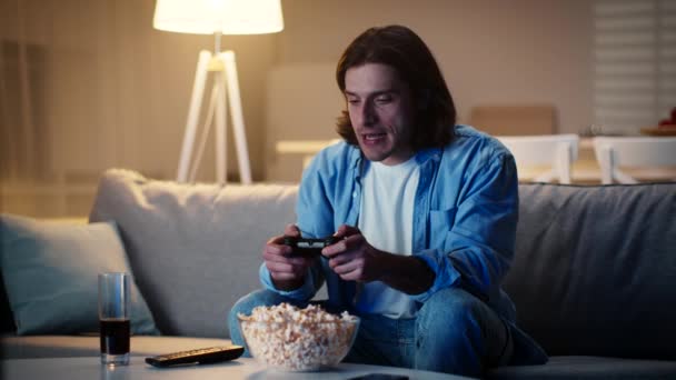 Giovane uomo che gioca ai videogiochi in serata, vincendo e celebrando la sua vittoria, seduto da solo sul divano di casa - Filmati, video