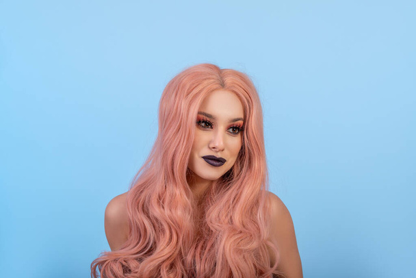 Portrait of cheerful young woman in pink long wig. Long eyelashes and black lipstick. Blue background - Foto, imagen