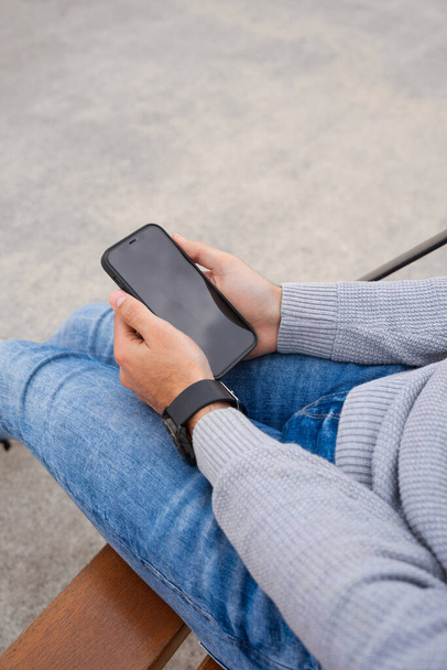 Накладний знімок рук, що тримає мобільний телефон, сидить на лавці. Він одягнений в наручний годинник, сині джинси і сірий светр. Він тримає мобільний телефон обома руками
. - Фото, зображення