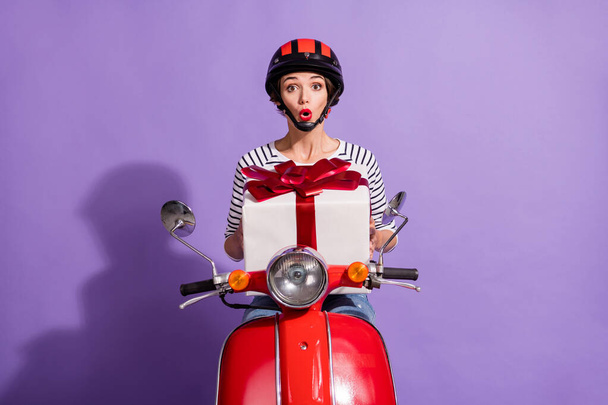 Portrait photo of shocked girl sitting on scooter keeping big present box opened mouth helmet isolated on bright purple color background - Foto, afbeelding