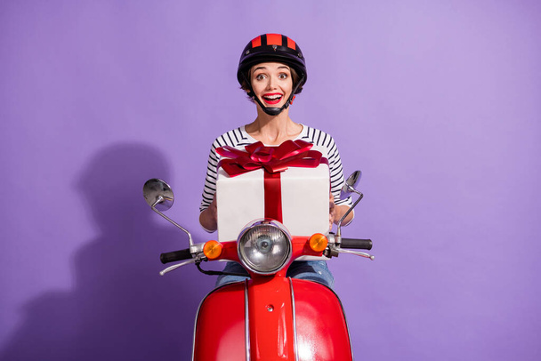 Portrait photo of cheerful female delivery worker driving bike giving wrapped gift isolated on vivid violet color background - Valokuva, kuva