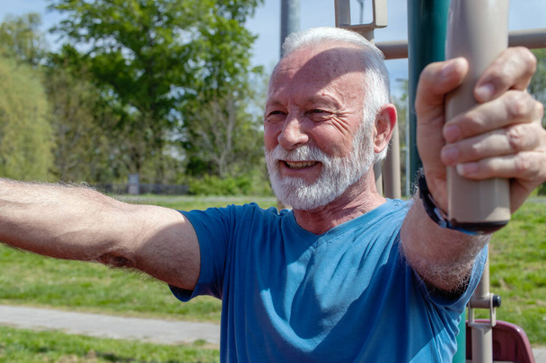 Starszy siwy mężczyzna z brodą ćwiczy na publicznej sali gimnastycznej na świeżym powietrzu.  - Zdjęcie, obraz