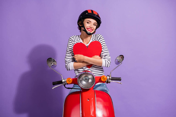 Portrait photo of happy girlfriend sitting on bike embracing red heart smiling wearing helmet isolated on vibrant purple color background - 写真・画像