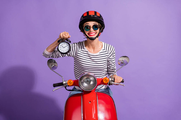 Portrait photo of girl in helmet on driving bike showing clock delivery in time smiling isolated on vibrant violet background - Φωτογραφία, εικόνα