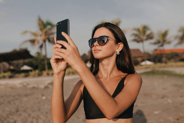 Stylish lovely woman with dark hair in sunglasses taking a picture on smartphone on the beach with palm trees on background. High quality photo - Photo, Image