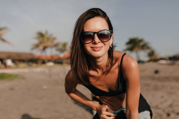 Imádnivaló lány rövid sötét frizurával napszemüvegben pózol a kamera előtt az egzotikus homokos parton. Kiváló minőségű fénykép - Fotó, kép
