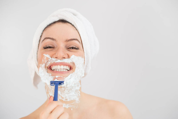 A woman with a white terry towel on her head and with shaving foam on her face shaves on a white background - Foto, afbeelding