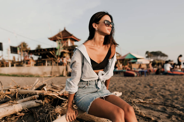 Adorabile bella ragazza con i capelli corti scuri in gonna e camicia blu seduta sulla spiaggia sabbiosa alla luce del sole. Foto di alta qualità - Foto, immagini