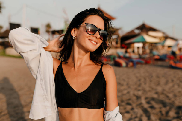 Retrato al aire libre de la encantadora chica linda con el pelo oscuro vistiendo top negro y camisa posando a la luz del sol en la playa de arena. Foto de alta calidad - Foto, imagen