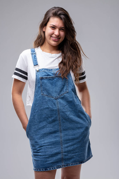 Confident casual trendy young woman in a denim pinafore standing in a relaxed pose with hands in pockets giving the camera a lovely friendly smile - Fotografie, Obrázek
