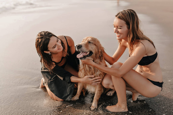 Dos chicas guapas en la playa cerca del océano jugando con un perro al atardecer. Foto de alta calidad - Foto, Imagen