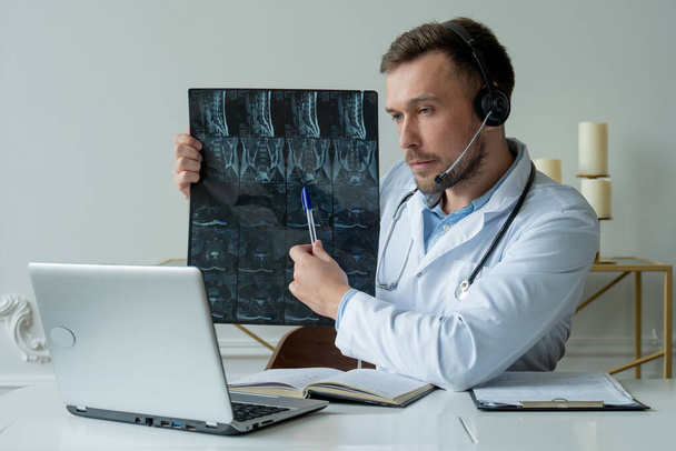 Male doctor listening to patient during telemedicine session. Man doctor having online consultation on laptop holding x-ray image and discussing diagnosis - Fotó, kép
