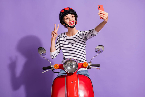 Photo of lady ride scooter take selfie telephone show v-sign wear helmet striped shirt isolated purple color background - Foto, imagen