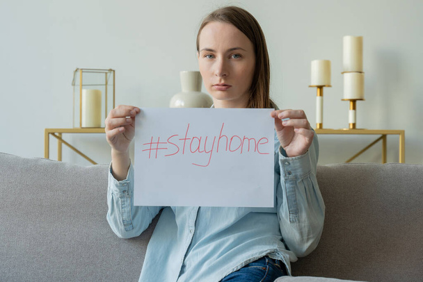 A woman holding a piece of paper with the inscription STAY HOME is sitting on the sofa - Foto, Imagen
