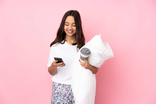 Young woman in pajamas isolated on pink background holding coffee to take away and a mobile - Foto, afbeelding