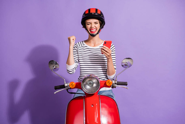 Photo of crazy girl ride motorbike raise fist hold telephone wear helmet striped shirt isolated purple color background - Фото, изображение