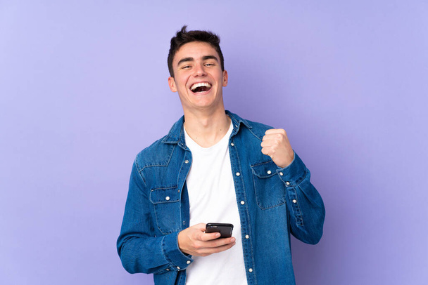 Teenager kavkazský pohledný muž izolované na fialovém pozadí s telefonem ve vítězné pozici - Fotografie, Obrázek