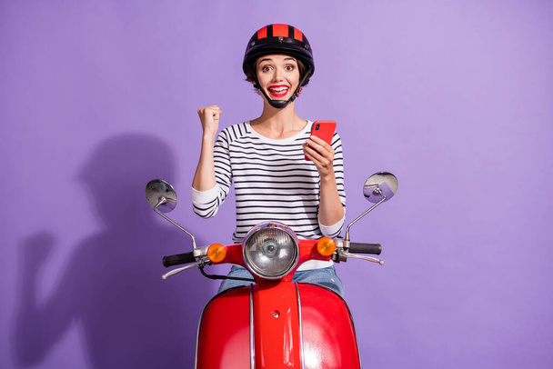 Photo of lady ride moped raise fist hold smartphone wear helmet striped shirt isolated violet color background - Valokuva, kuva