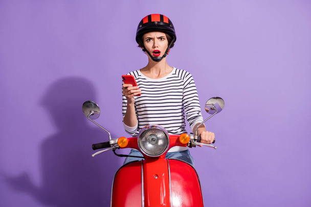 Photo of confused upset lady ride scooter hold telephone open mouth wear helmet striped shirt isolated violet color background - Foto, imagen