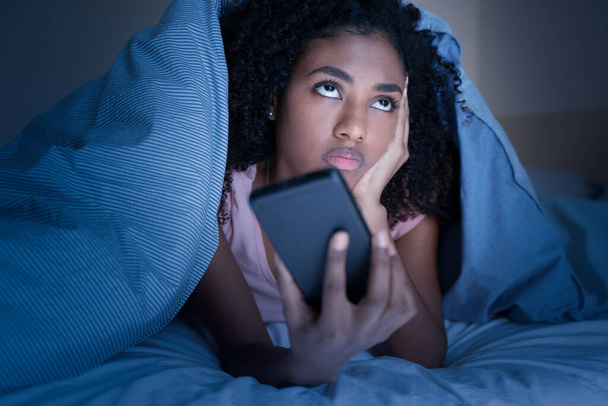 Сонная женщина лежит в постели под одеялом, используя смартфон ночью. - Фото, изображение