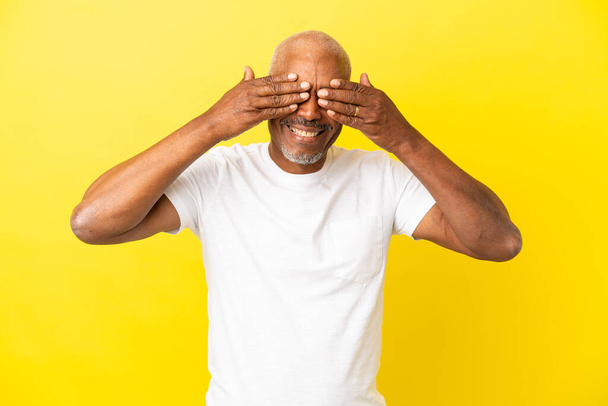 Kubański Senior odizolowany na żółtym tle okrywający oczy dłońmi i uśmiechnięty - Zdjęcie, obraz