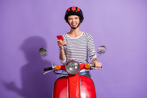 Photo of excited crazy lady drive retro bike hold cellphone close eyes wear helmet striped shirt isolated purple color background - Foto, Bild