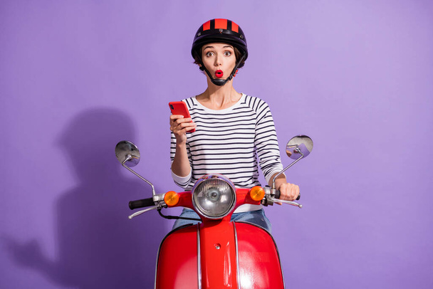 Photo of shocked lady drive motorcycle hold mobile phone open mouth wear helmet striped shirt isolated purple color background - Фото, изображение