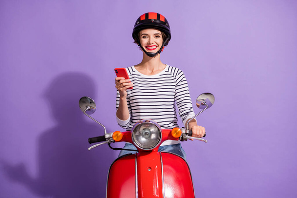Foto von positiven Mädchen fahren Moped halten Telefon tragen Helm gestreiftes Hemd isoliert violette Farbe Hintergrund - Foto, Bild