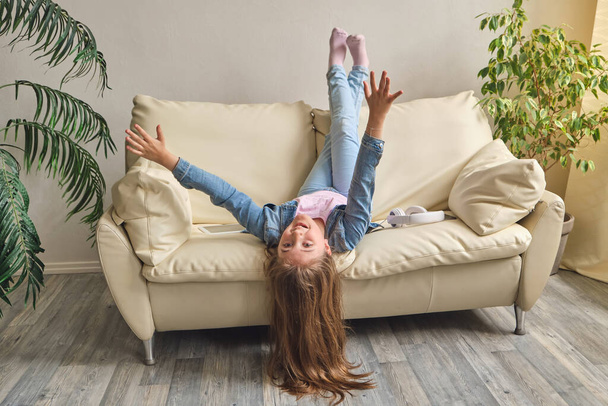 Красивая маленькая девочка лежит вверх ногами на диване, смотрит в камеру и улыбается, играя дома. - Фото, изображение