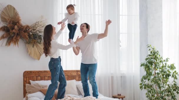 Young family Caucasian parents with small daughter playing at home on bed, strong father man holding baby infant little girl with one hand acrobatic pose mom catches child laughing throws kid into air - Footage, Video