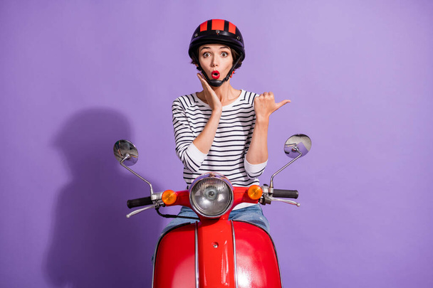 Photo of excited girl ride motorbike indicate thumb empty space palm cheek wear helmet striped shirt isolated purple color background - 写真・画像